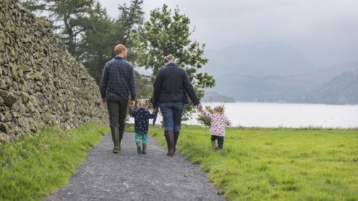 A family of four, two adults and two young children, hold hands as they walk down a path towards the lakeshore, against a mountain backdrop